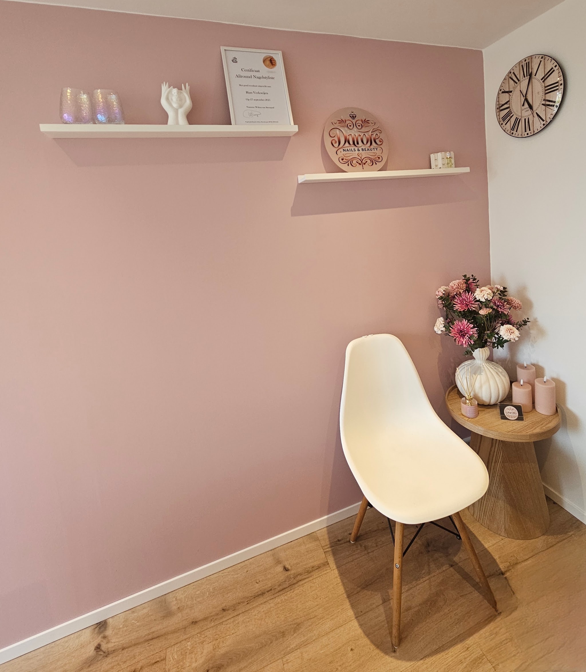 Roze muurhoek met witte wandplanken, het ingelijste logo, een klok, bloemen en een witte stoel